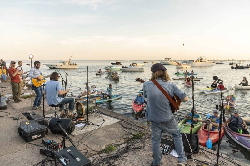 People in kayaks look on from the water as a band plays music on a pier
